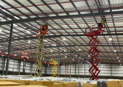 Electricians on scissor lifts fitting high-bay lighting in a warehouse